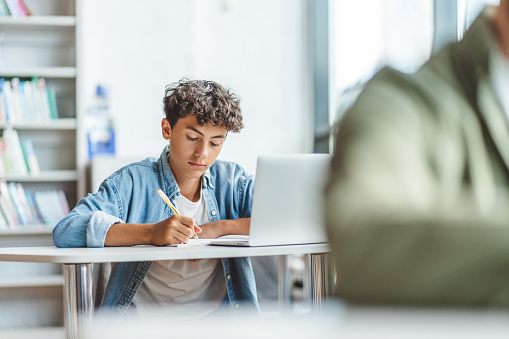 Pensive, smart schoolboy taking notes learning languages, preparing for exam using laptop in modern classroom. Serious brunette guy wearing blue shirt doing homework. Back to school, education concept
