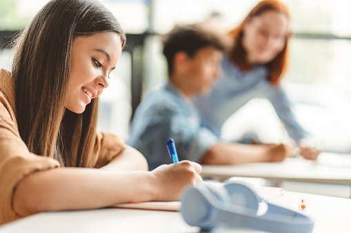 Beautiful, smiling schoolgirl wearing stylish casual clothes sitting at desk, taking notes, holding pen, learning language, exam preparation. Attractive teenage girl in classroom doing homework