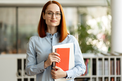 Portrait of successful middle aged businesswoman, manager looking at camera. Smart confident university student wearing eyeglasses holding laptop, book standing in modern library, education concept