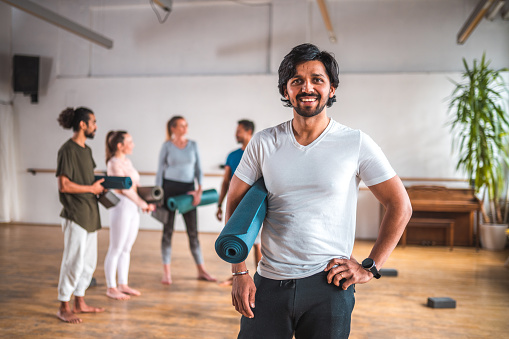 Portrait of an Indian man after sports training, posing for the camera standing in a room with other students of pilates holding mats in blurred background. Looking at the camera.
