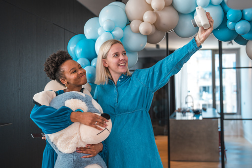 Two young, radiant friends, one pregnant and Caucasian, the other African-American with curly hair, take a cheerful selfie. They are holding a Polaroid camera and a stuffed toy, enjoying a joyous moment at a baby shower celebration.
