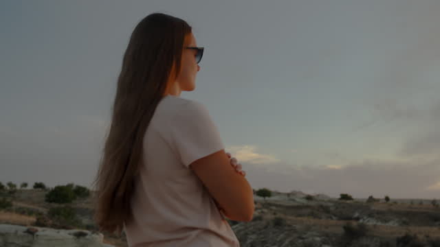 A lonely young woman stands with folded hands and watches the setting sun at the edge of the cliff.