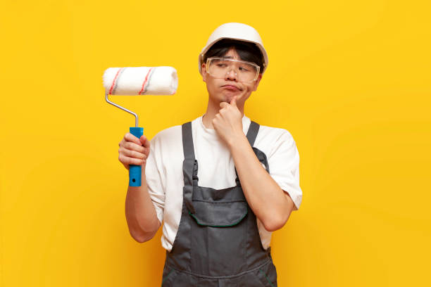 istock pensive asian guy painter in uniform holds roller and thinks plans on yellow isolated background 1764141868