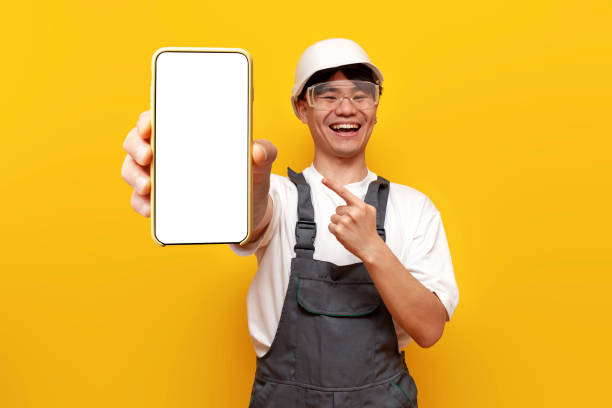 istock Asian guy builder in uniform and safety glasses shows blank smartphone screen on yellow isolated background 1764141872