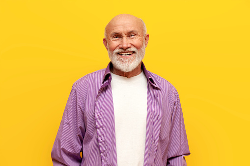 old bald grandfather with gray beard in purple shirt smiling on yellow isolated background, elderly pensioner looking at the camera