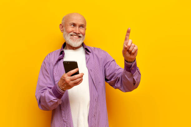 istock old bald grandfather with gray beard uses smartphone and points with his finger on yellow isolated background 1764144012