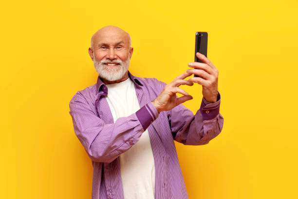 istock old bald grandfather with gray beard uses smartphone and selects online on yellow isolated background 1764144532