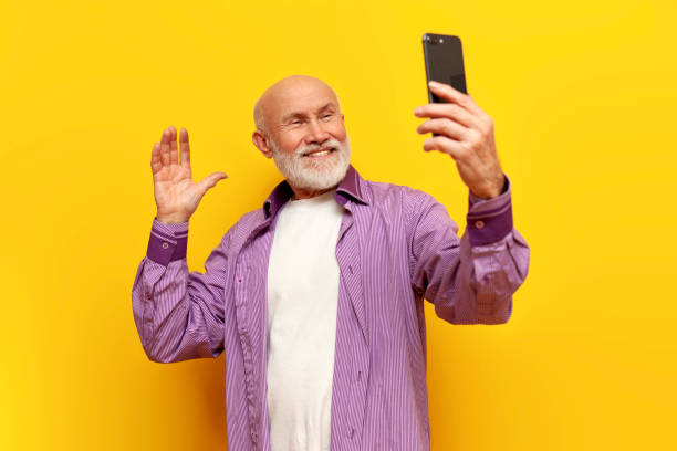 istock old bald grandfather with gray beard uses smartphone and greets on yellow isolated background 1764144536