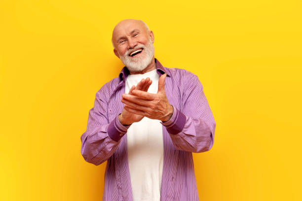 old bald grandfather with gray beard applauds and laughs on yellow isolated background, elderly pensioner