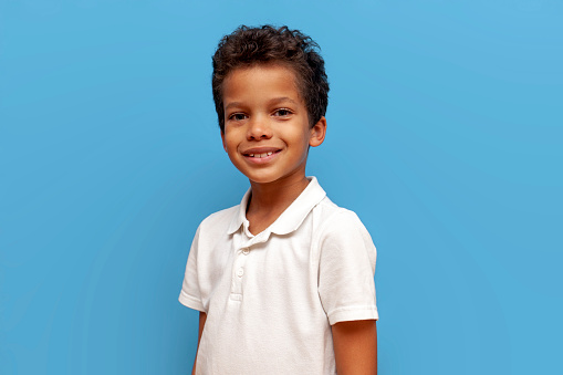 African American teenage boy in white t-shirt looking at camera on blue isolated background, curly schoolboy smiling
