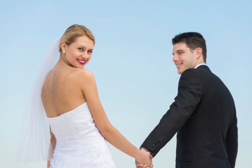 Portrait of happy young bride holding groom's hand against clear blue sky