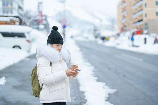 woman using mobile phone and drinking hot Coffee or Tea with snow in winter season during travel in Niseko. landmark and popular for attractions in Hokkaido, Japan. Travel and Vacation concepts