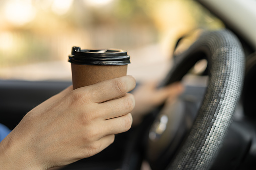 Female hand holding paper coffee cup behind the wheel. Takeaway drink in car. Side view
