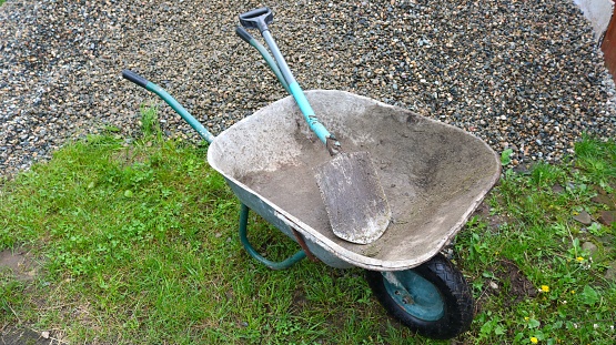 empty garden cart with a metal body and a shovel lying in it next to a pile of gray gravel on a green lawn top view, gardening equipment for use in cargo transportation, moving stone in a wheelbarrow