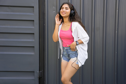Young beautiful Indian teenager girl wearing headphones listening to music from mobile phone, Dancing and singing. outdoor. Summer season.