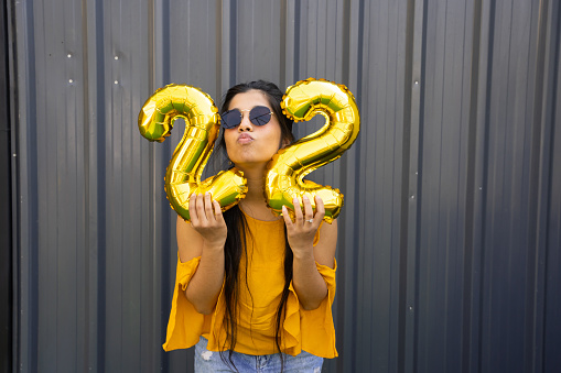 Cheerful beautiful young indian woman wearing casual cloths and sunglasses holding golden balloon number twenty two.