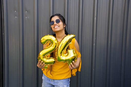 Cheerful beautiful young indian girl wearing casual cloths and sunglasses holding golden balloon number twenty four.