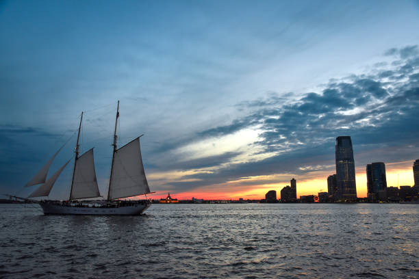 夕暮れ時にハドソン川を航行するスクーナー船とジャージーシティのスカイラインを背景に - 米国、ニューヨーク市