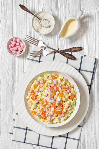Ambrosia Salad with pineapple, mandarin oranges, yogurt, mini marshmallows, coconut and whipped cream in white bowl on white wooden table with ingredients, american recipe, vertical view