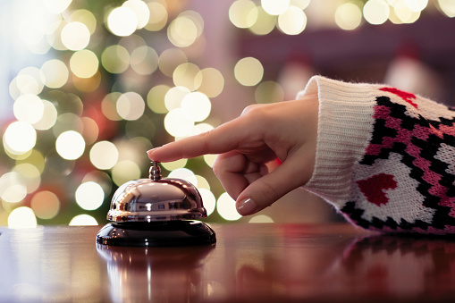 Christmas travel. Hand of guest ringing bell on reception desk of guesthouse and color shining garland on christmas tree on background. Hotel, restaurant.