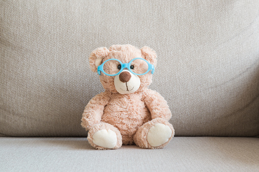 Brown teddy bear with eyeglasses with blue round frame sitting on sofa at home. Children vision concept. Closeup. Front view.