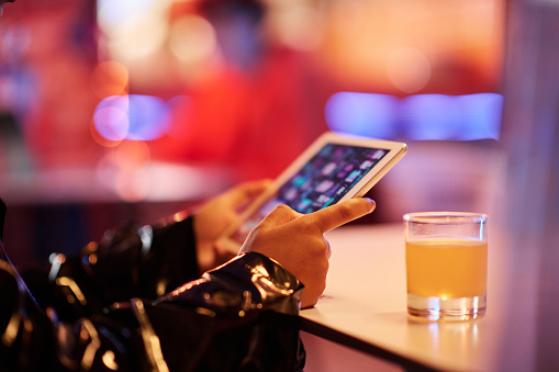 Focus on hands of young woman in black shiny jacket holding tablet while sitting by table in cyberpunk bar, having drink and playing video game