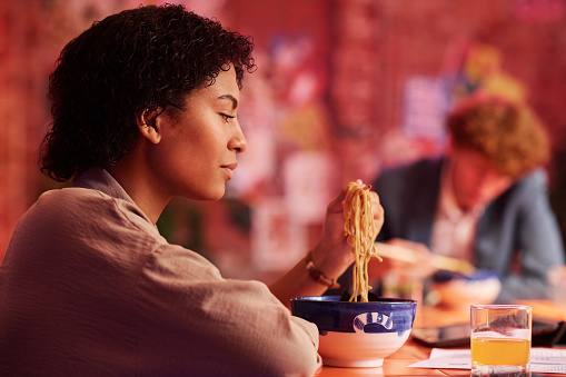 Side view of young hungry African American woman looking at appetizing ramen on chopsticks while enjoying lunch in Japanese cafe or bar