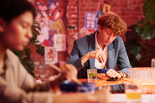 Focus on young businessman in formalwear eating ramen with chopsticks while sitting by table in Japanese cafe or bar with cyberpunk design