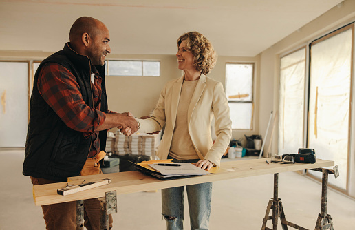 Contractor shakes hands with a female homeowner, signifying an agreement and partnership. They are indoors, surrounded by documents, suggesting a business deal for a home improvement project.