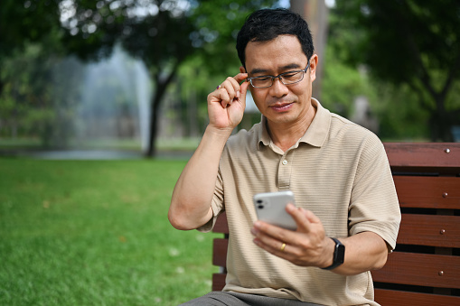 Smiling middle age man wearing glasses using mobile phone in the public park.