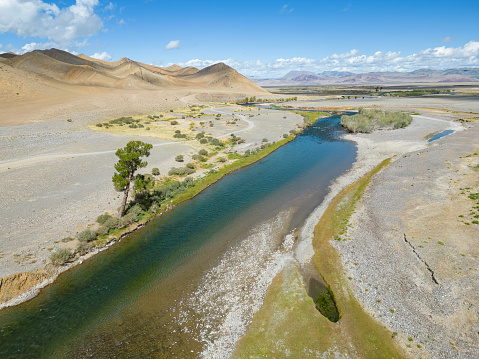 An aerial drone view of a freshwater river running through barren landscape in the Bayan Ulgii province of the Altai Mountains in Western Mongolia. The remote location is home to Kazakh nomadic herders who graze their livestock in the mountainous region during the summer months.