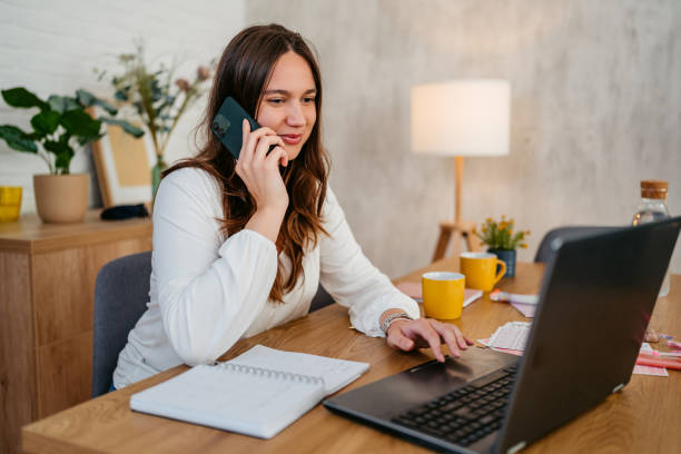 istock Young Woman Talking On The Phone While Working On A Laptop At The Kitchen Table 1764691881