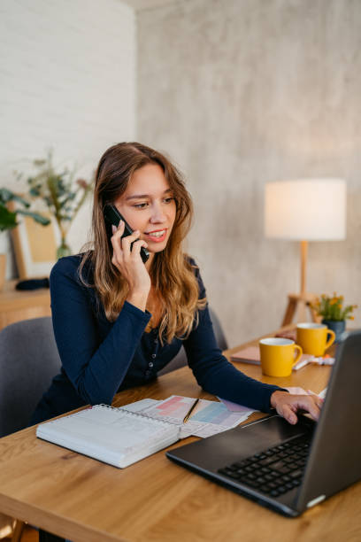 istock Young Woman Talking On The Phone While Working On A Laptop At The Kitchen Table 1764695789