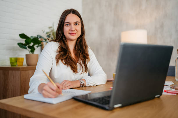 istock Young Woman Writing In A Notebook While Working On A Laptop At The Kitchen Table 1764696333