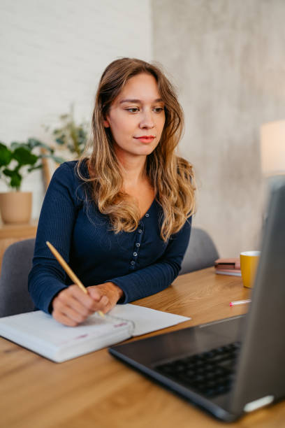 istock Young Woman Writing In A Notebook While Working On A Laptop At The Kitchen Table 1764696338