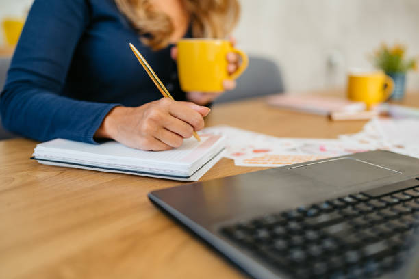 istock Young Woman Writing In A Notebook While Working On A Laptop At The Kitchen Table 1764696853