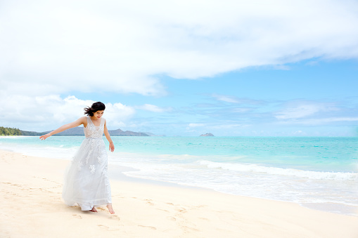 Teen girl in white dress walking barefoot on hawaiian beach by blue Pacific Ocean