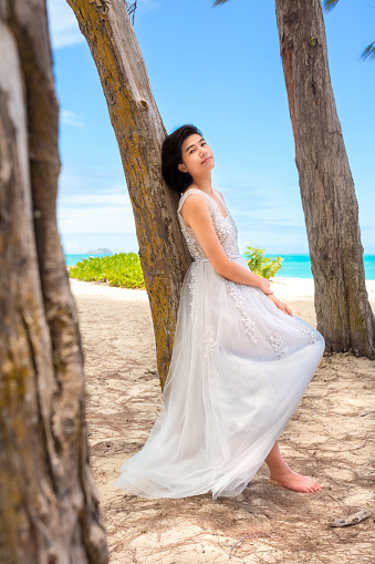 Teen girl in white dress resting against tall  trees along Hawaiian beach, blue ocean in background