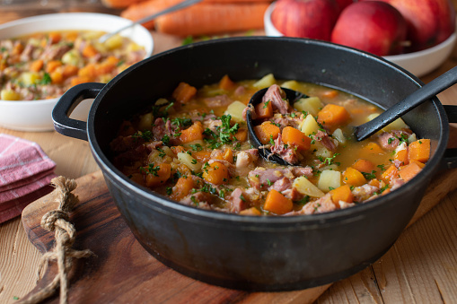 Delicious autumn or winter stew with carrots, potatoes and smoked pork meat. Served ready to eat in a pot with ladle on rustic and wooden table background. Closeup, front view