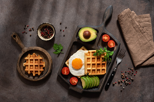 Healthy breakfast with waffles, fried egg, avocado and tomatoes on a dark background with sunlight and shadows. Top view, flat lay.