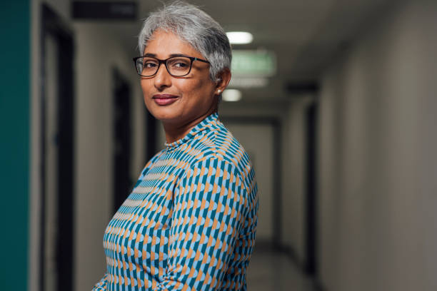 Proud of Her Work A portrait of a mid adult female doctor, standing in a low lit corridor in a hospital in Newcastle upon Tyne, North East England. She has her body facing away from the camera with her head turned to look at the camera as she has a contented look on her face. doctor general practitioner stock pictures, royalty-free photos & images