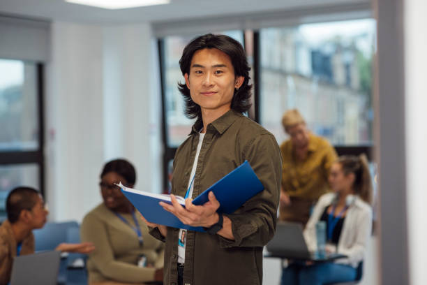 In His First Year of Medical Studies A male student nurse standing in an education classroom in a hospital in Newcastle upon Tyne, North East England. He is holding an open writing book while looking at the camera and smiling with other students behind him in the background, out of focus. They are all studying nursing. male chinese student stock pictures, royalty-free photos & images
