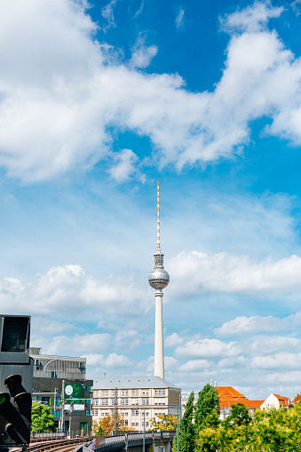 The Fernsehturm Tower is seen against a blue sky with white clouds. The photo is taken from a distance, with buildings and trees in the foreground.