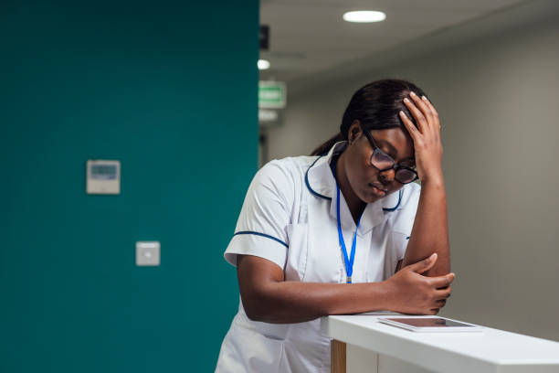 An Overworked Medical Student A portrait of a young female student nurse, standing in a reception area in a hospital in Newcastle upon Tyne, North East England. She is leaning on the reception desk with her elbow and head in her hand while looking down at a digital tablet with a concerned look on her face, she is using the tablet to educate her while she works. melancholy computer support specialist stock pictures, royalty-free photos & images