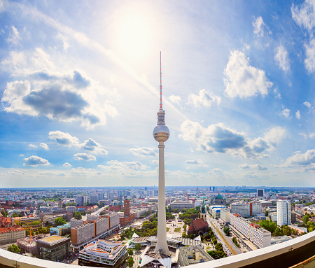 A panoramic photo of the Berliner Fernsehturm, a television tower in Berlin, Germany. The tower is located in the center of the image and is the tallest structure in the photo. The photo was taken from a high vantage point. The photo was taken on a sunny day with blue skies and scattered clouds. The background consists of a cityscape with buildings, roads, and trees.