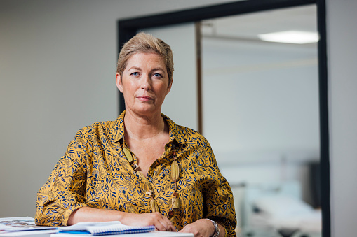 A portrait of a mid adult female doctor, standing in a reception area in a hospital in Newcastle upon Tyne, North East England. She is leaning on the reception desk while looking at the camera with a contented look on her face.