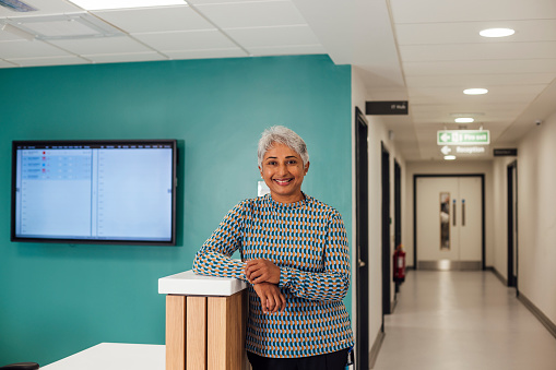 A portrait of a mid adult female doctor, standing in a reception area in a hospital in Newcastle upon Tyne, North East England. She is leaning on the reception desk while looking at the camera and smiling.