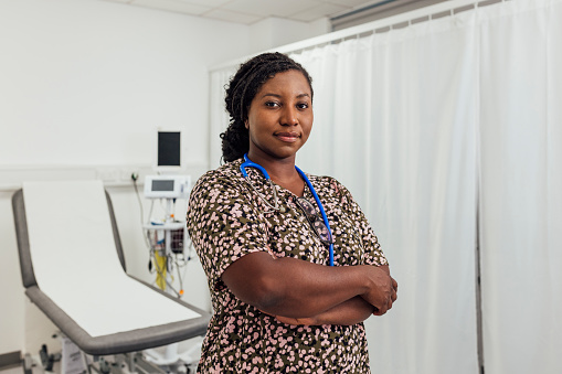 A portrait of a mid adult female doctor wearing a stethoscope, standing on a ward in a hospital in Newcastle upon Tyne, North East England. She is looking at the camera with her arms crossed and a contented look on her face. There is examination bed behind her, ready for her next patient.