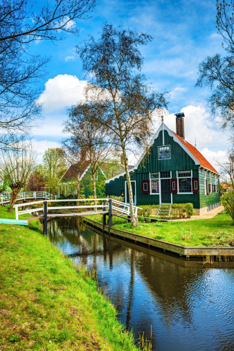 One beautiful Dutch scene with traditional wooden houses by the canal. Visible are many green trees, bridges and beautiful cloudscape over the reflection in the canal. Zaanse Schans, Netherlands.