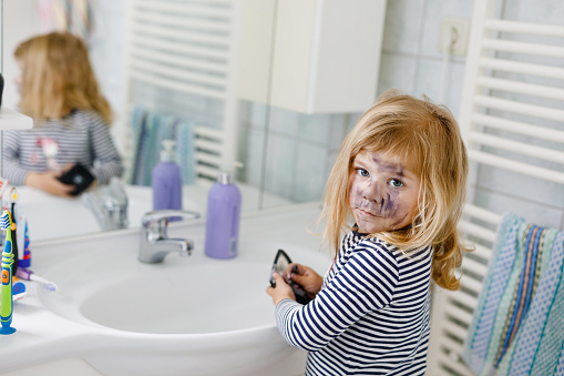 Funny little toddler girl using mother's make up and painting face with eye shadows. Happy baby child making experiments with cosmetics of mother. Kid around.